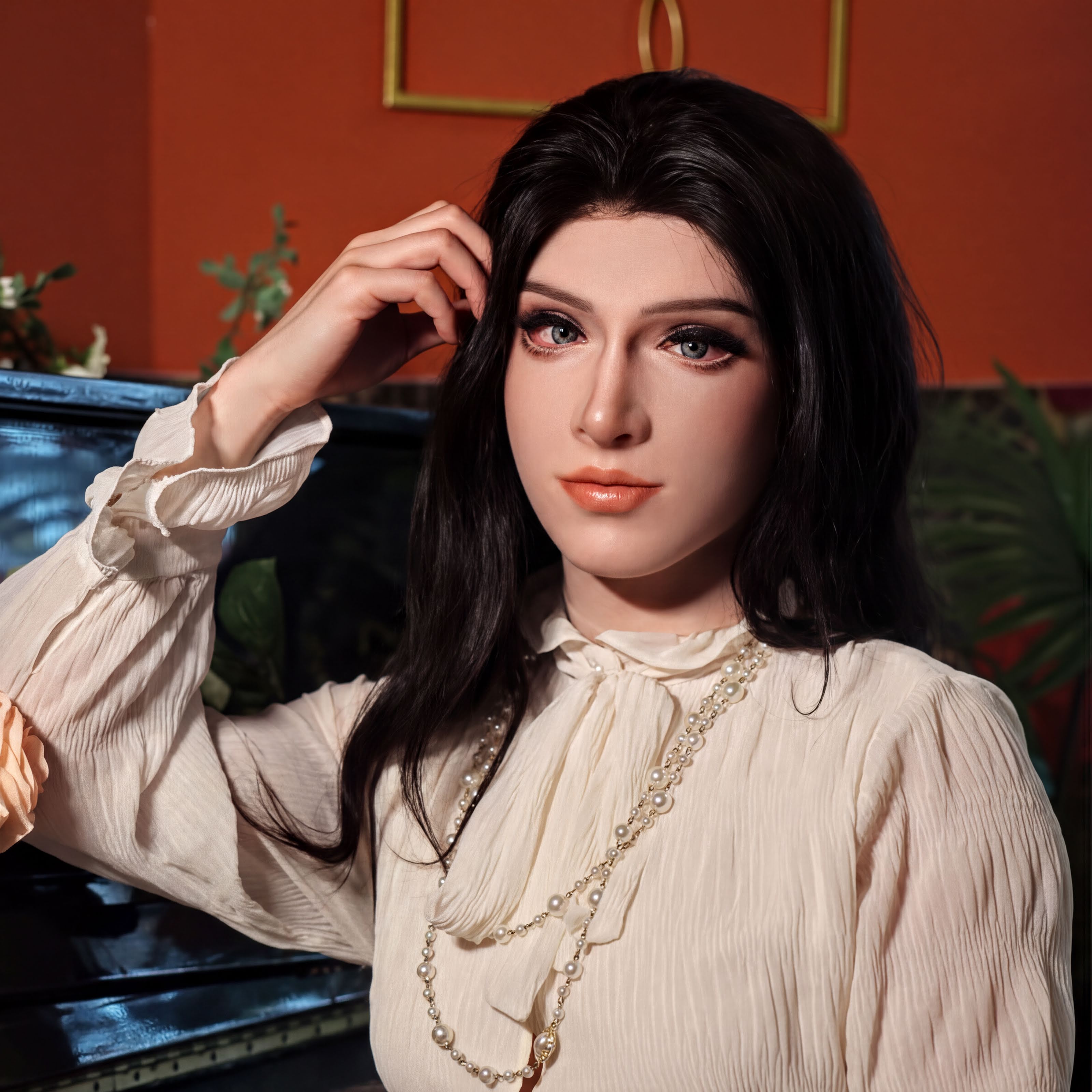 A woman with long dark hair wearing a white blouse and pearls poses in front of a piano, with a potted plant visible in the background.
