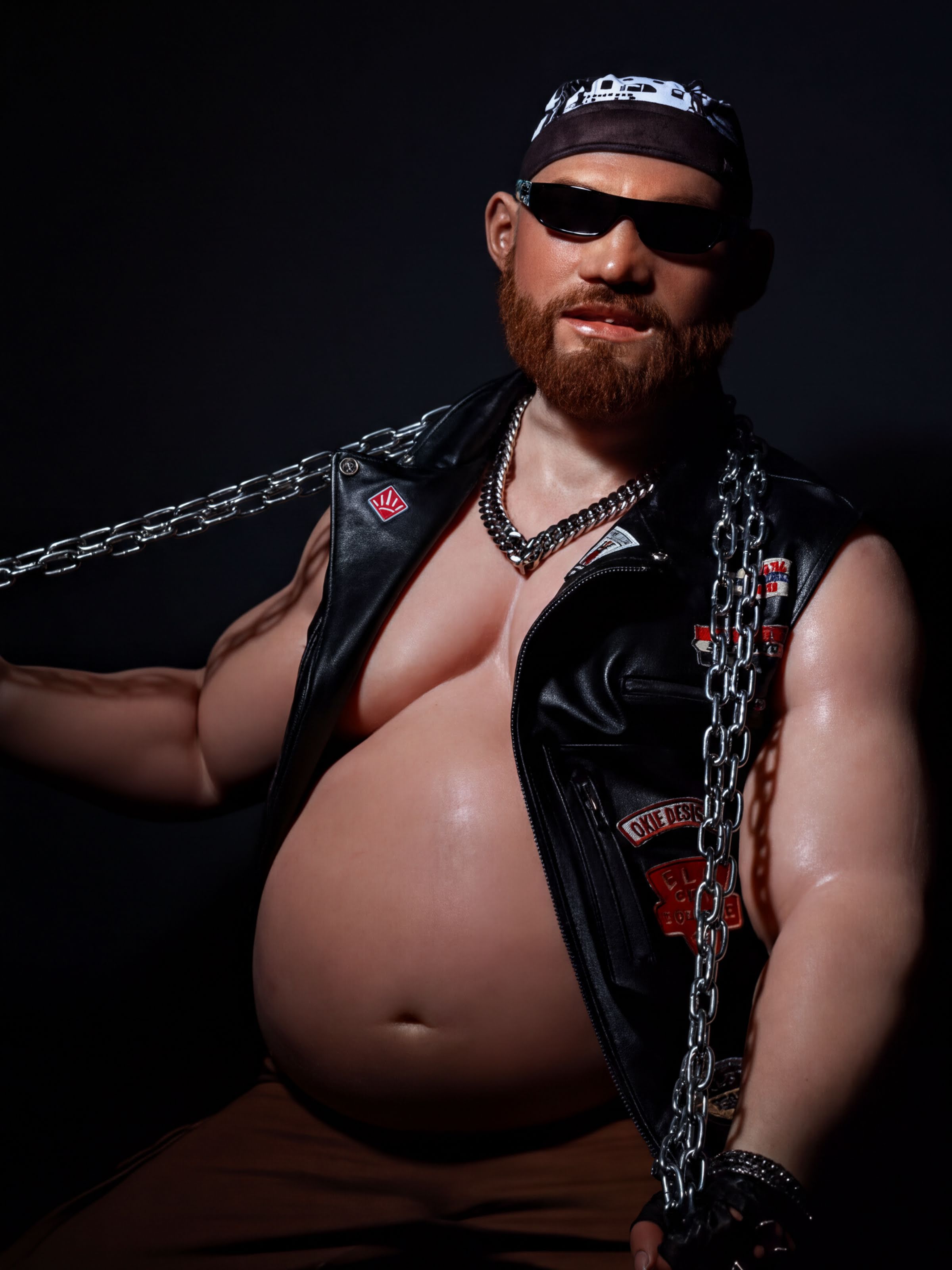 A muscular man with a beard wearing a leather jacket, sunglasses, and holding a chain, posing in a dark setting.