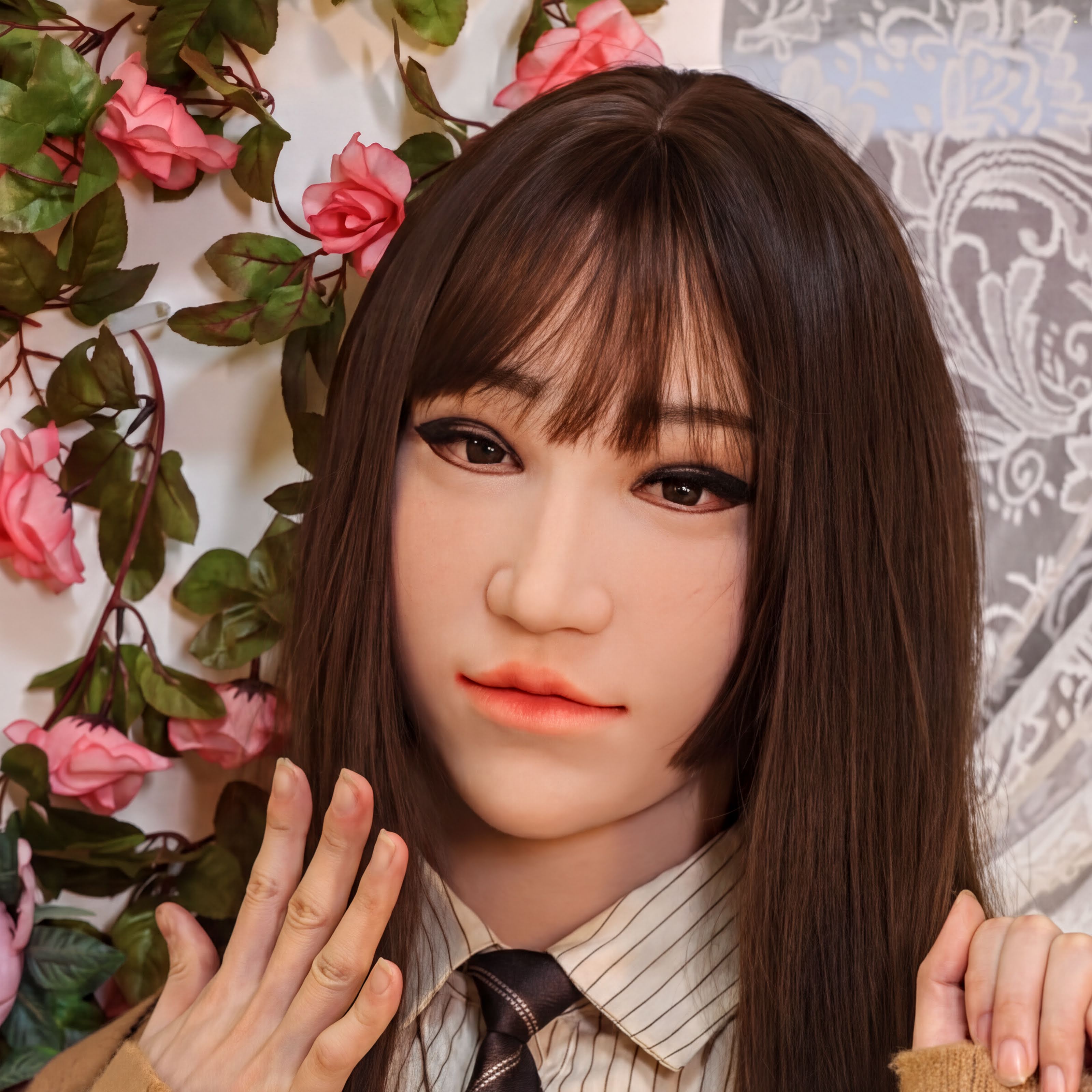 A close-up of a person wearing a mask, with long brown hair and a serious expression. The background features pink flowers and a white curtain.