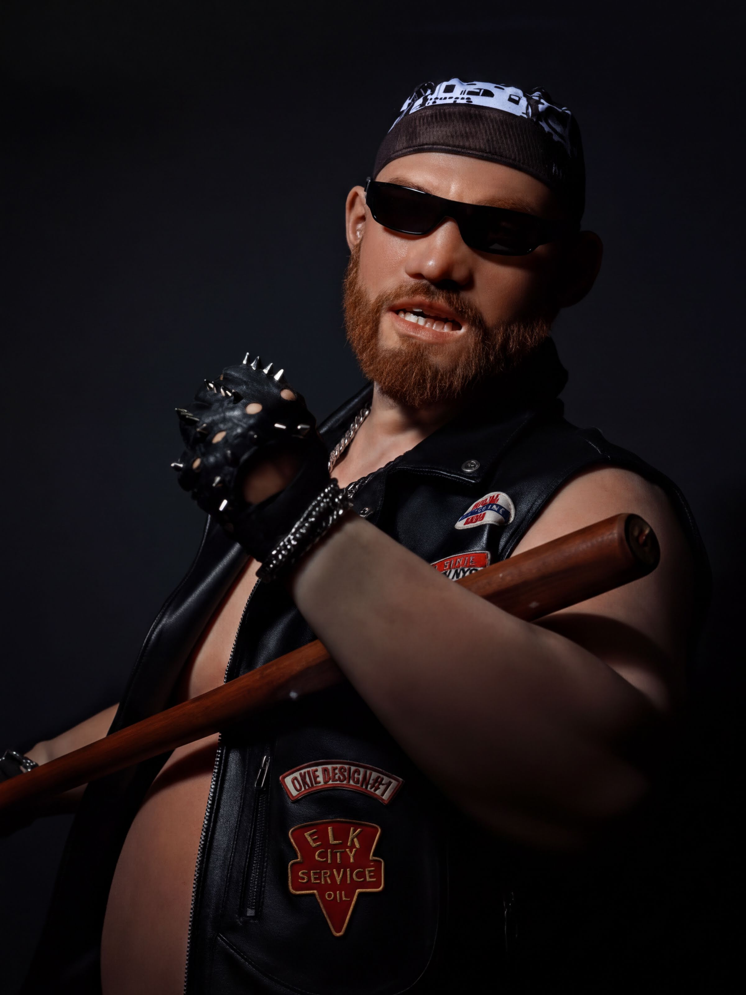 A bearded man wearing sunglasses, a leather jacket, and a bandana is holding a wooden stick or pipe in a dark setting.