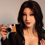 A woman with dark hair and makeup is holding a camera in the image.