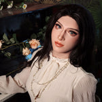 A woman with long dark hair and makeup is sitting at a piano, wearing a white blouse and a necklace.