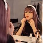 A woman with long, dark hair and a headband is sitting at a vanity, applying makeup in front of a mirror. She is wearing a black top and has makeup brushes and a bottle of lotion on the vanity.